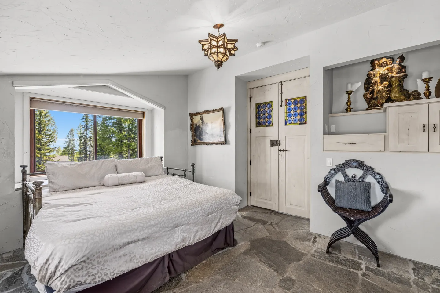 Bedroom with stone tile flooring, a metal-frame bed beside a picture window showing pine trees, built-in shelving, and an ornate pendant light fixture.