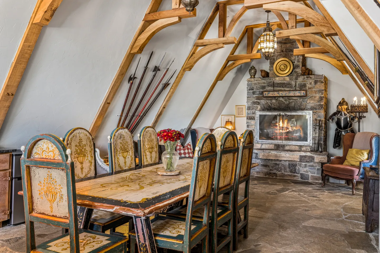 Dining area with an A-frame timber ceiling, long painted dining table, stone fireplace, and medieval-themed decor.