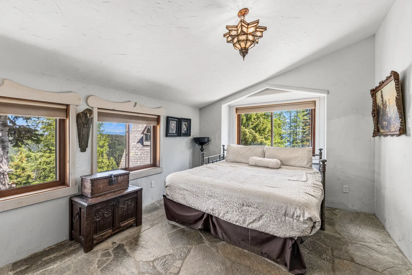 Bedroom with stone floor, light textured walls, a king-size bed, ornate hanging light, and windows showing trees and mountains.