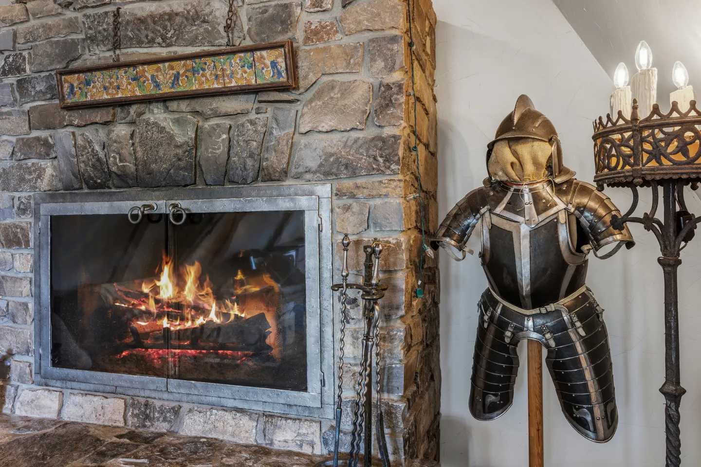 A lit stone fireplace with glass doors adjacent to a decorative suit of armor.