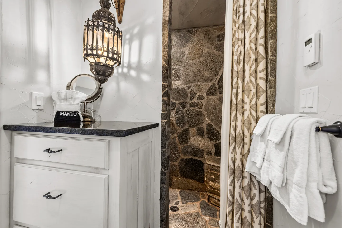 Bathroom with a white vanity, ornate lantern pendant light, and a stone-tiled walk-in shower partially behind a curtain.