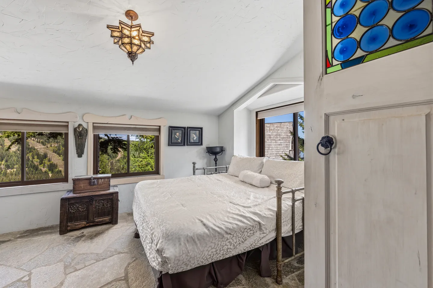 Bedroom viewed through a white doorway with stone-textured flooring, a metal-framed bed, and windows overlooking a wooded hillside.