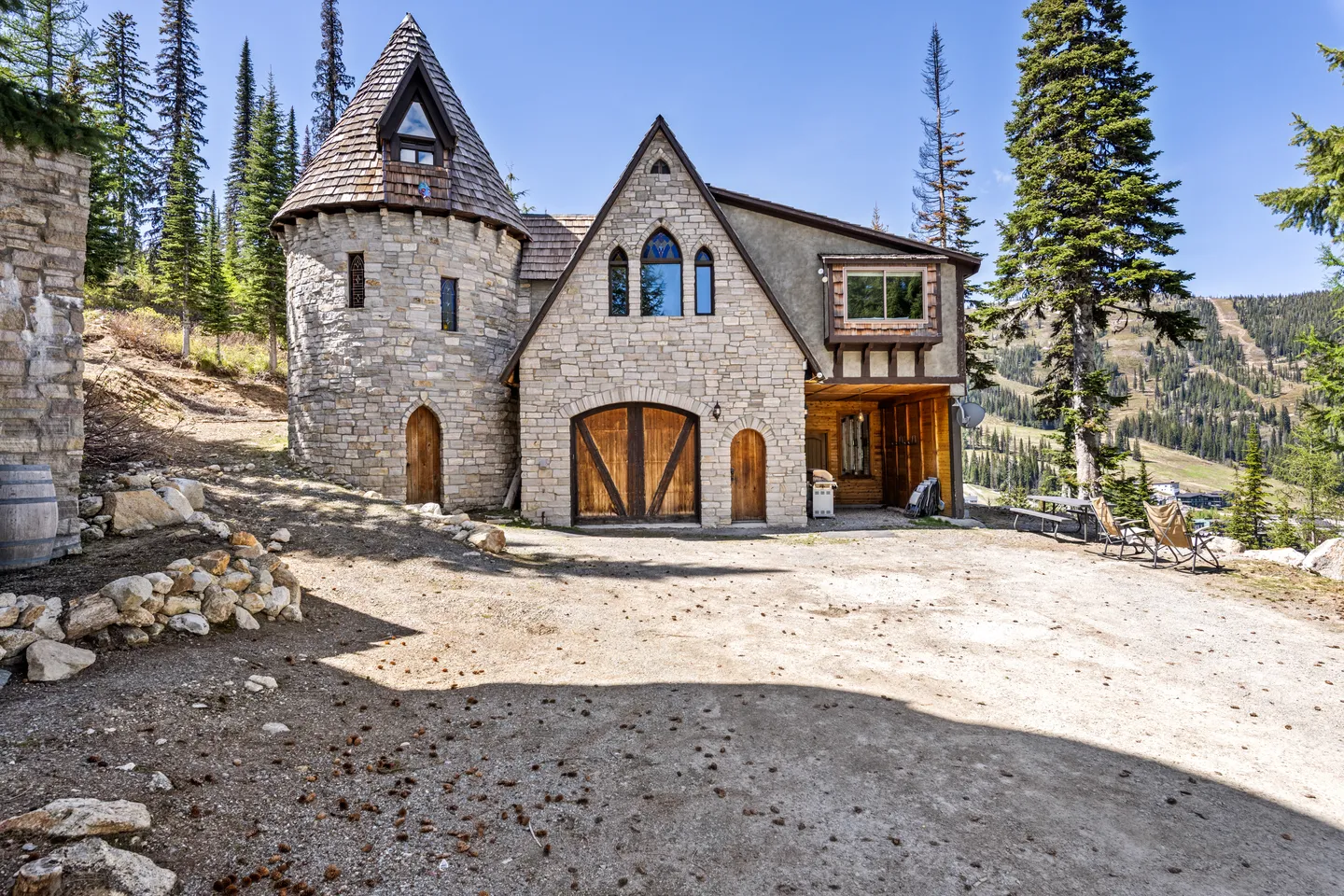 Exterior of a castle-style home featuring stone walls, a round tower with a conical roof, and arched wooden doors set against a mountainous forest backdrop.