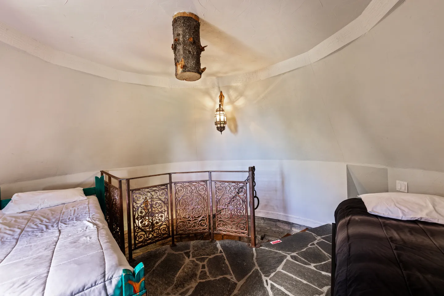 Loft bedroom inside a circular room with flagstone flooring, two twin beds, an ornate metal railing, and a rustic log ceiling fixture.