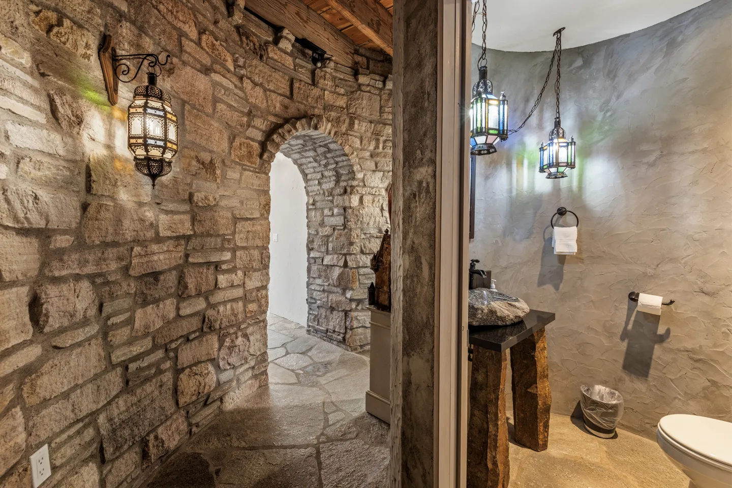 A stone hallway featuring an arched doorway and lantern sconce next to a bathroom with textured walls and hanging pendant lights.