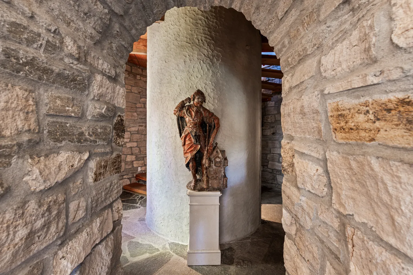 A stone archway looks through to a painted statue mounted on a pedestal in front of a curved textured pillar and stone floor.