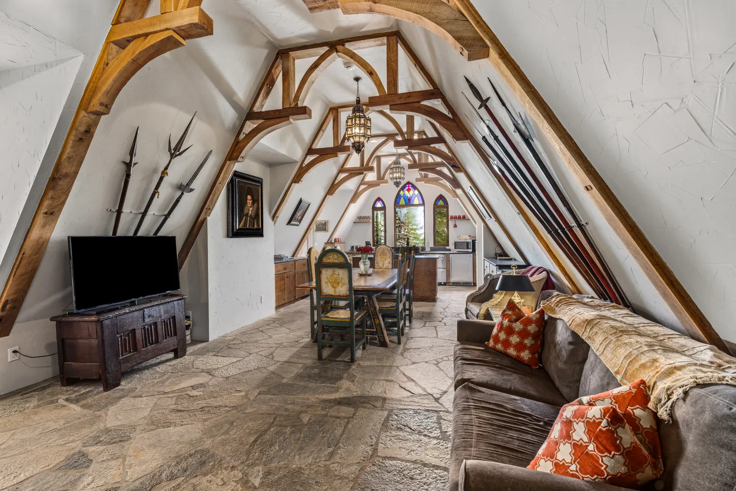 A long attic living space with exposed curved wood ceiling trusses, stone tile flooring, a couch, dining set, and medieval-style polearms on the wall.