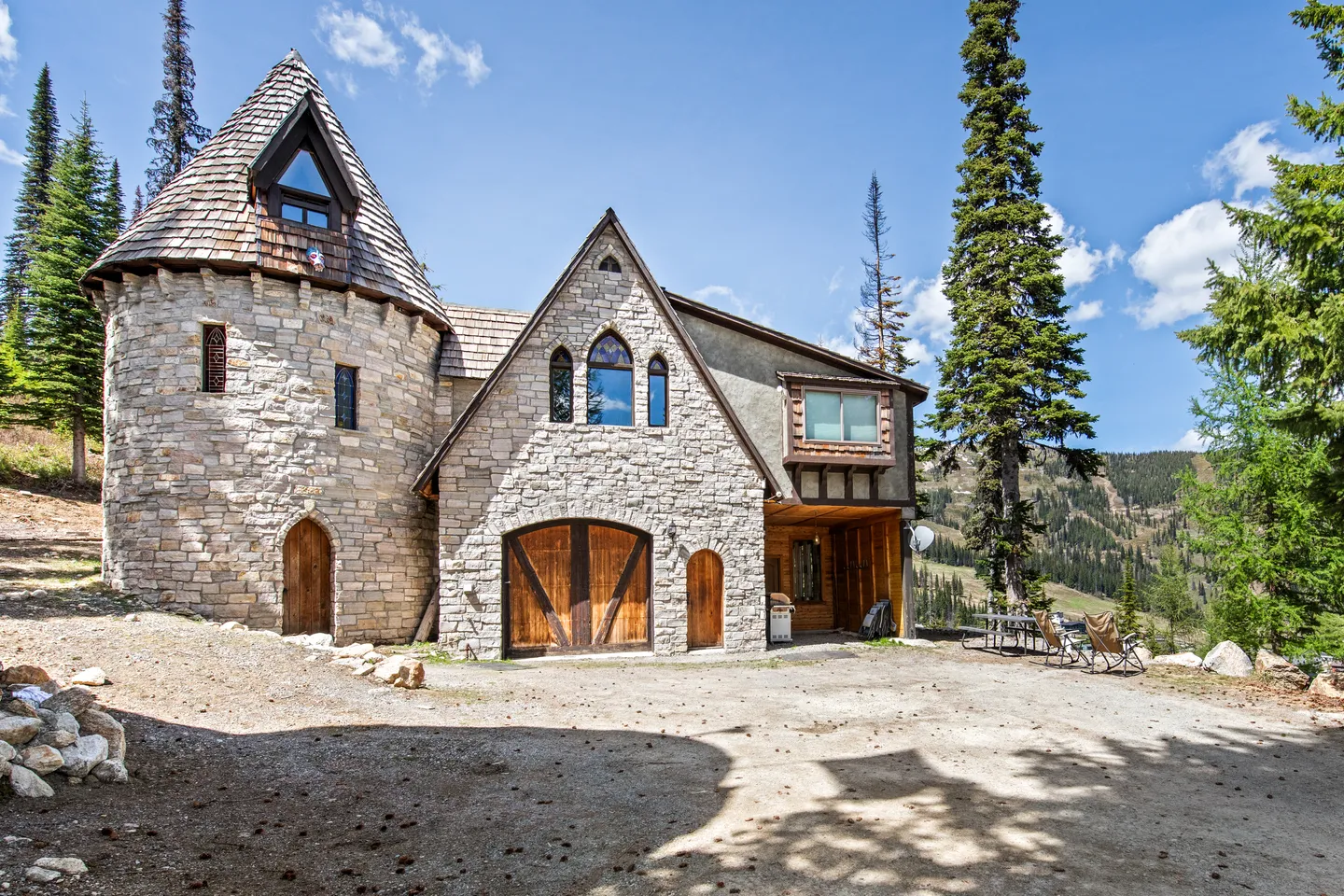 Exterior of a castle-style home with light stone masonry, a round turret, arched wooden doors, and pointed windows set in a wooded mountain area.