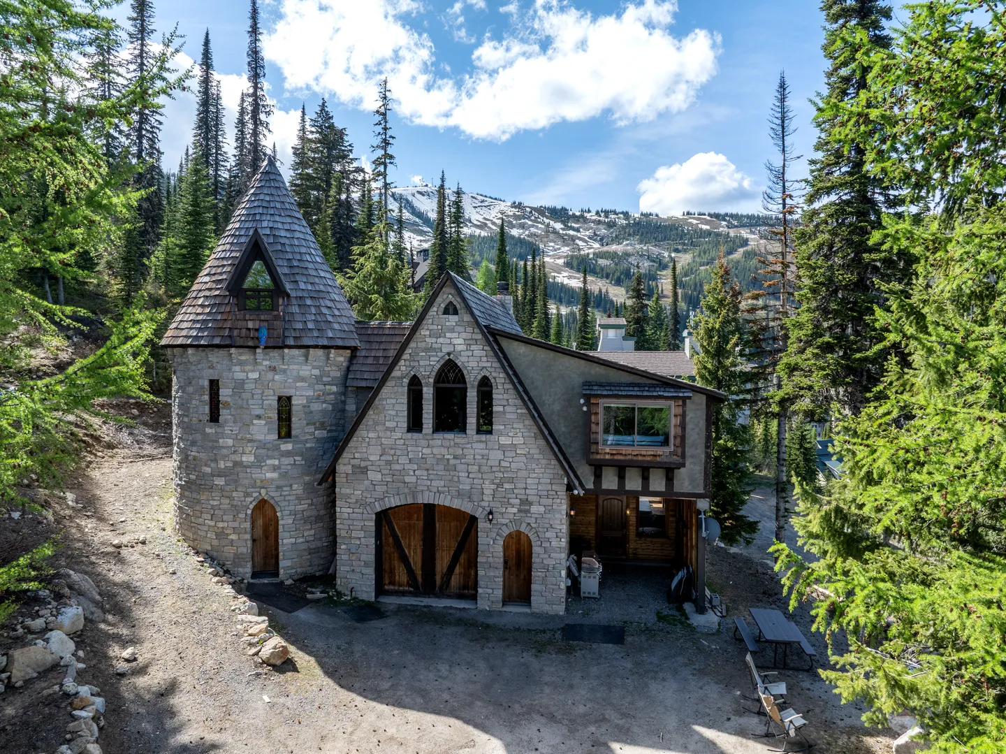 Exterior of a castle-style stone house with a round conical turret, arched doorways, and tall pine trees against a snowy mountain backdrop.