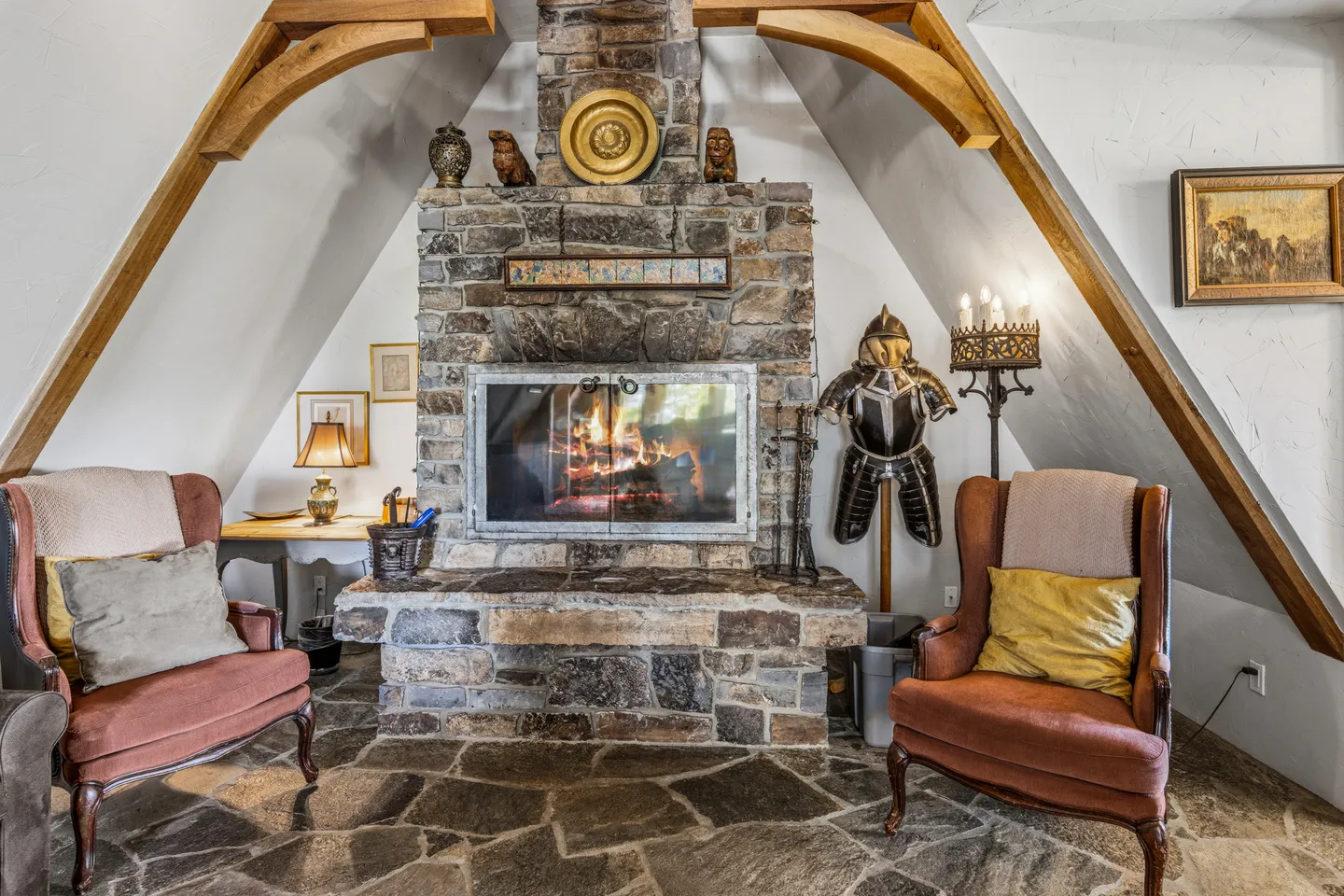 A stone fireplace with a lit fire flanked by two armchairs under sloped ceilings with exposed wooden corbels.