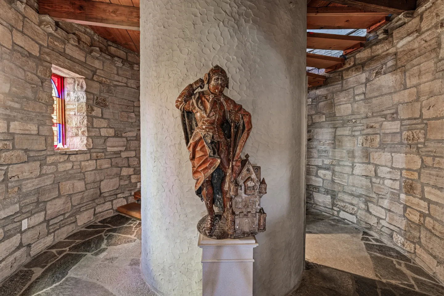 A stone tower stairwell featuring a central column with a sculpted knight figure on a pedestal, flagstone flooring, and exposed stone walls.