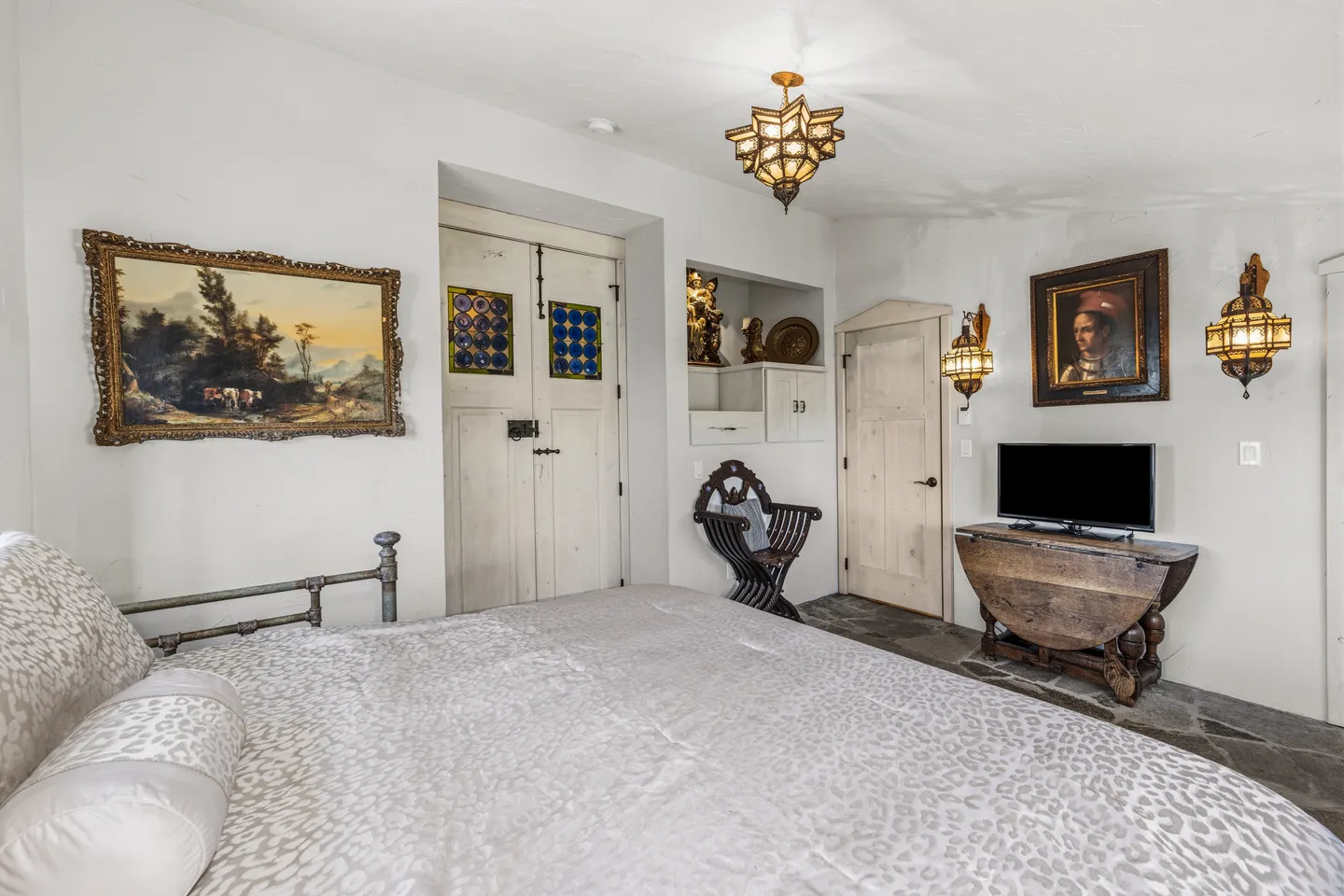 Bedroom with a patterned bedspread, stone flooring, decorative light fixtures, framed artwork, and a television on a drop-leaf table.