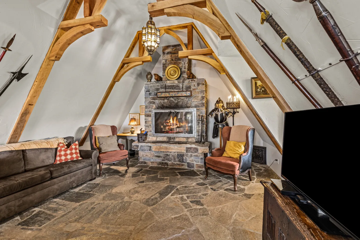 Living area under an A-frame ceiling with timber trusses, stone fireplace, stone flagstone floor, and medieval decor including polearms and a suit of armor.