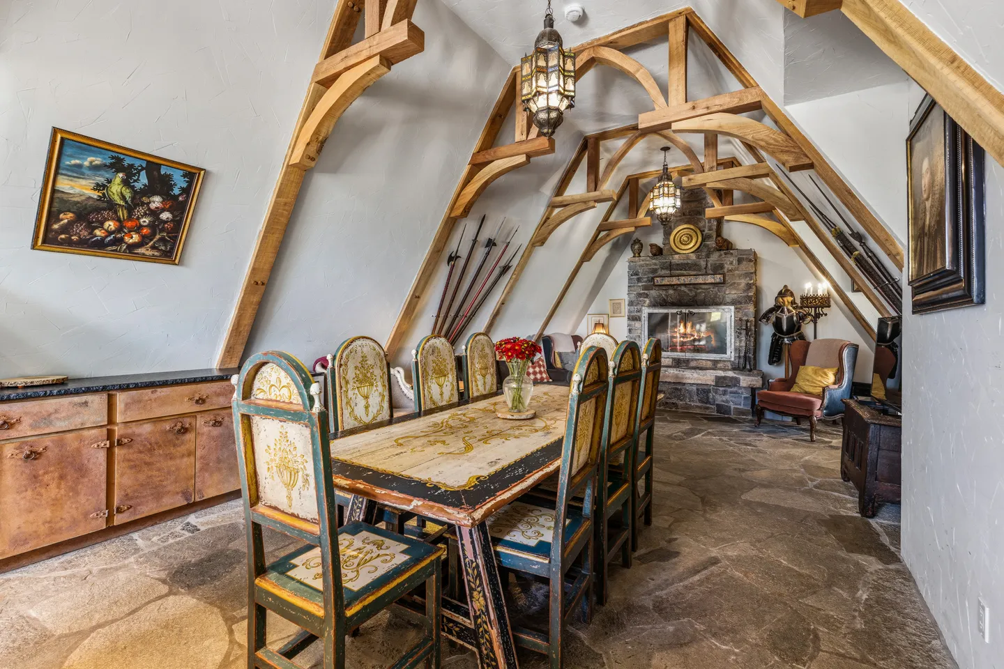 Dining and sitting room under an A-frame ceiling with arched timber beams, a long painted dining table, and a stone fireplace.