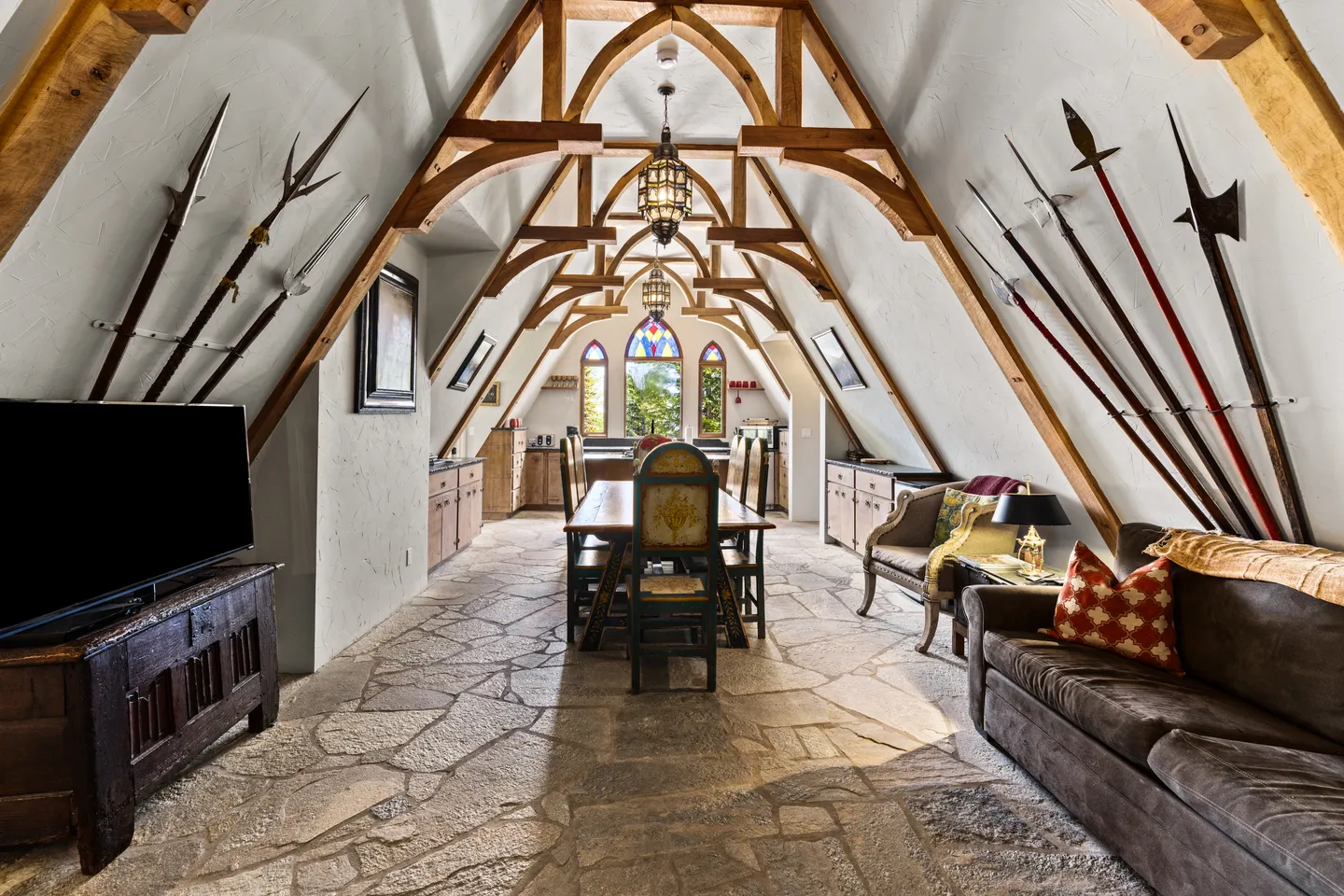 Medieval-themed long room with vaulted timber beam ceilings, stone floor, dining table, seating, and polearms mounted on the sloped walls.