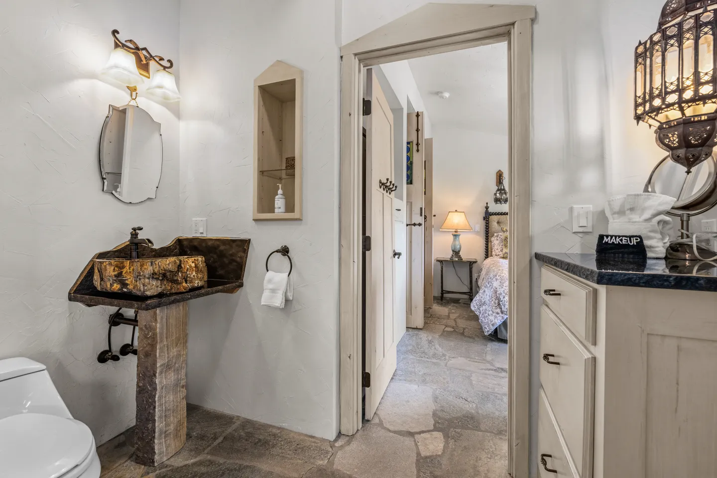 Bathroom with stone-flagged floor, rustic stone-look pedestal sink, and open doorway leading to a bedroom.