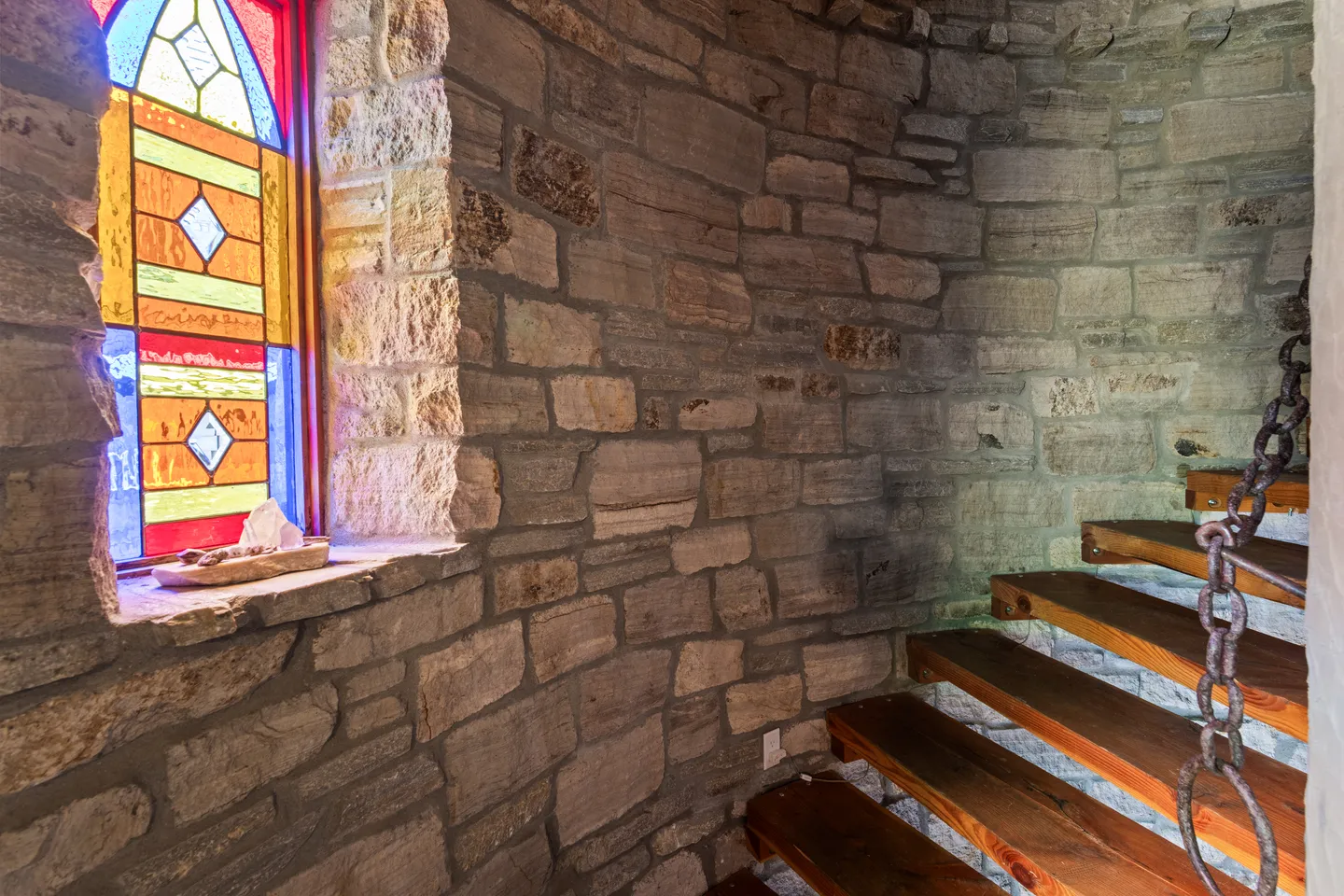 A curved stone stairwell featuring floating wooden steps, a heavy metal chain handrail, and a stained glass window.