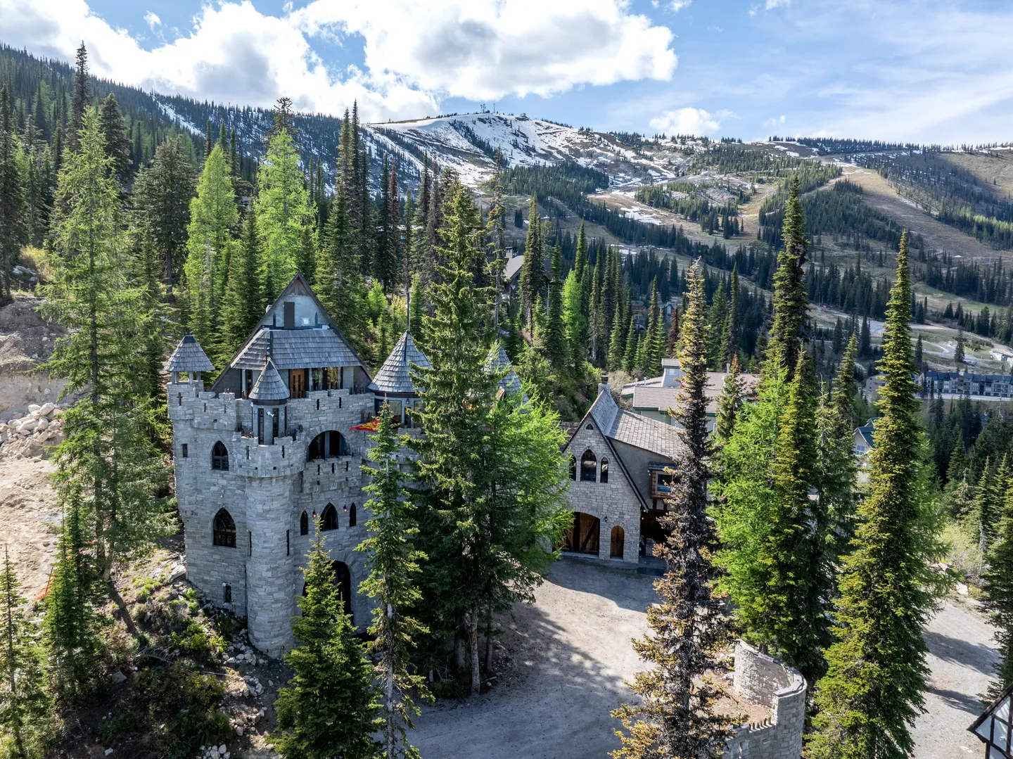 Aerial view of a stone castle-style estate surrounded by evergreen trees with snow-dusted mountains in the background.