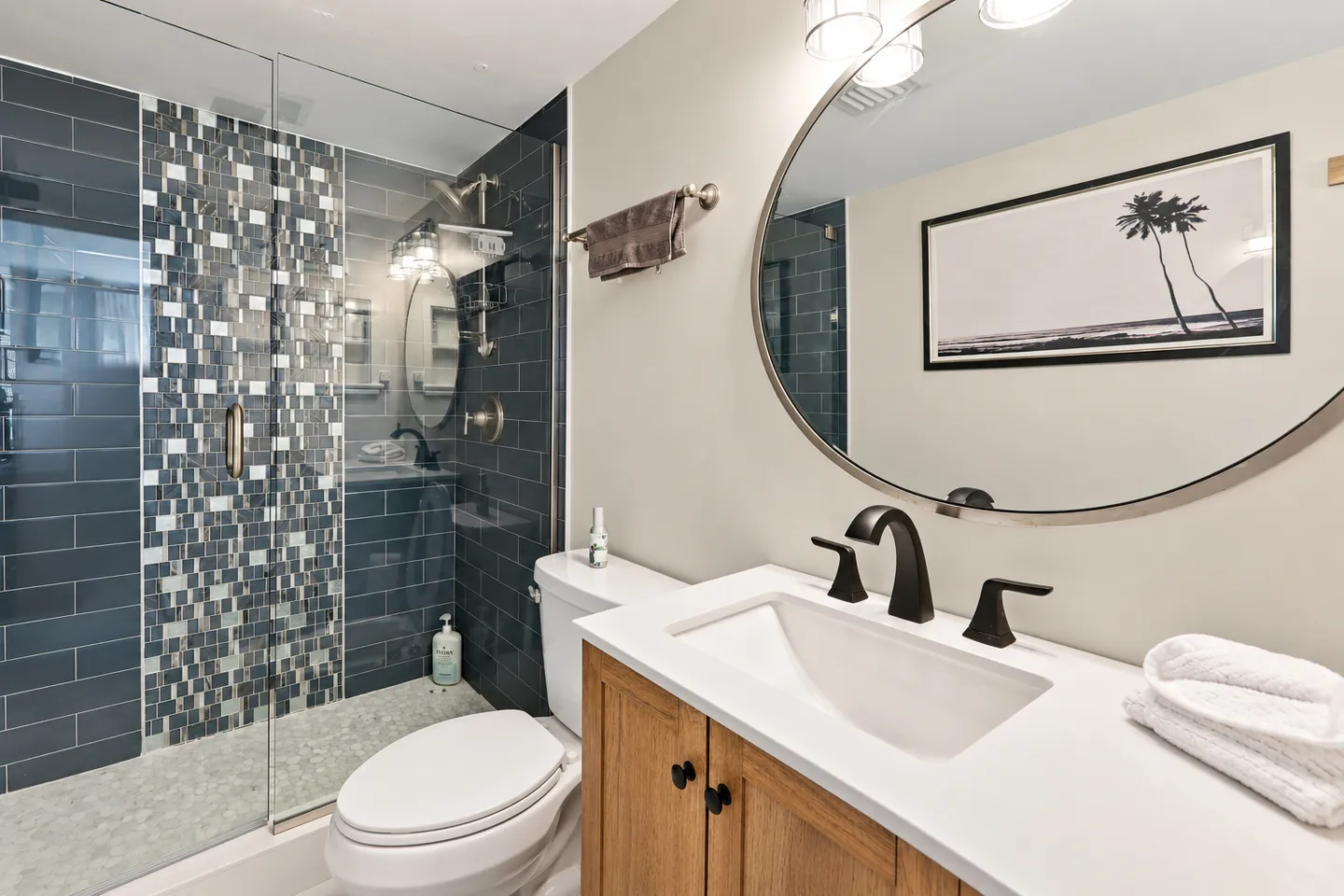 Bathroom with a wood vanity, white countertop, oval mirror, toilet, and walk-in shower with dark and mosaic tile.