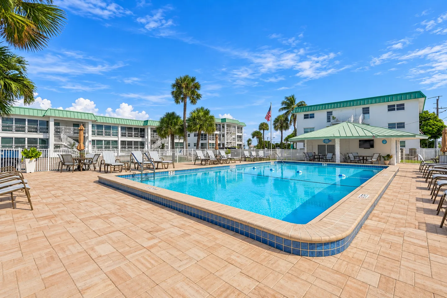 Outdoor swimming pool surrounded by a paved patio, lounge chairs, and apartment buildings under a sunny blue sky with palm trees.