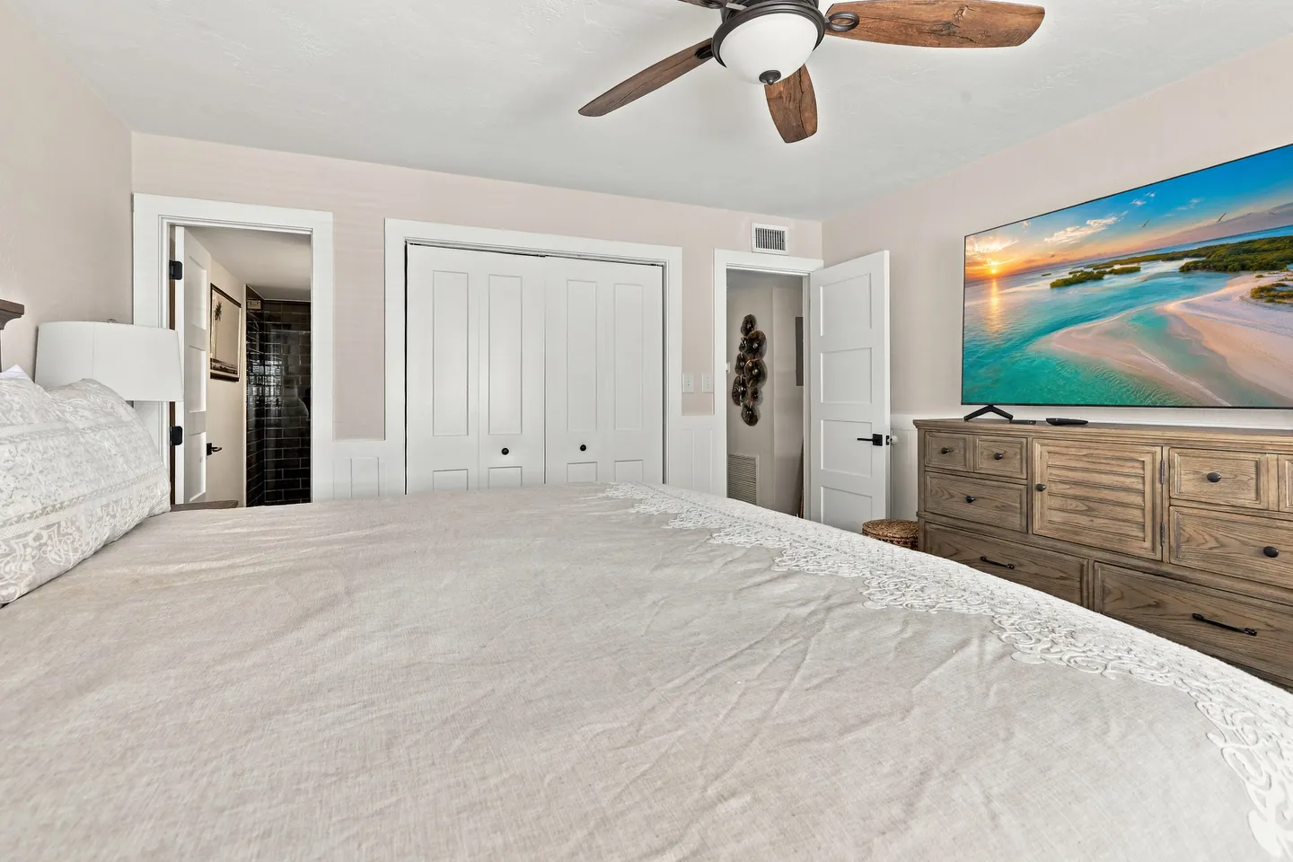 Bedroom featuring a made bed in the foreground, wooden dresser with a mounted television, ceiling fan, and doors leading to a closet and bathroom.