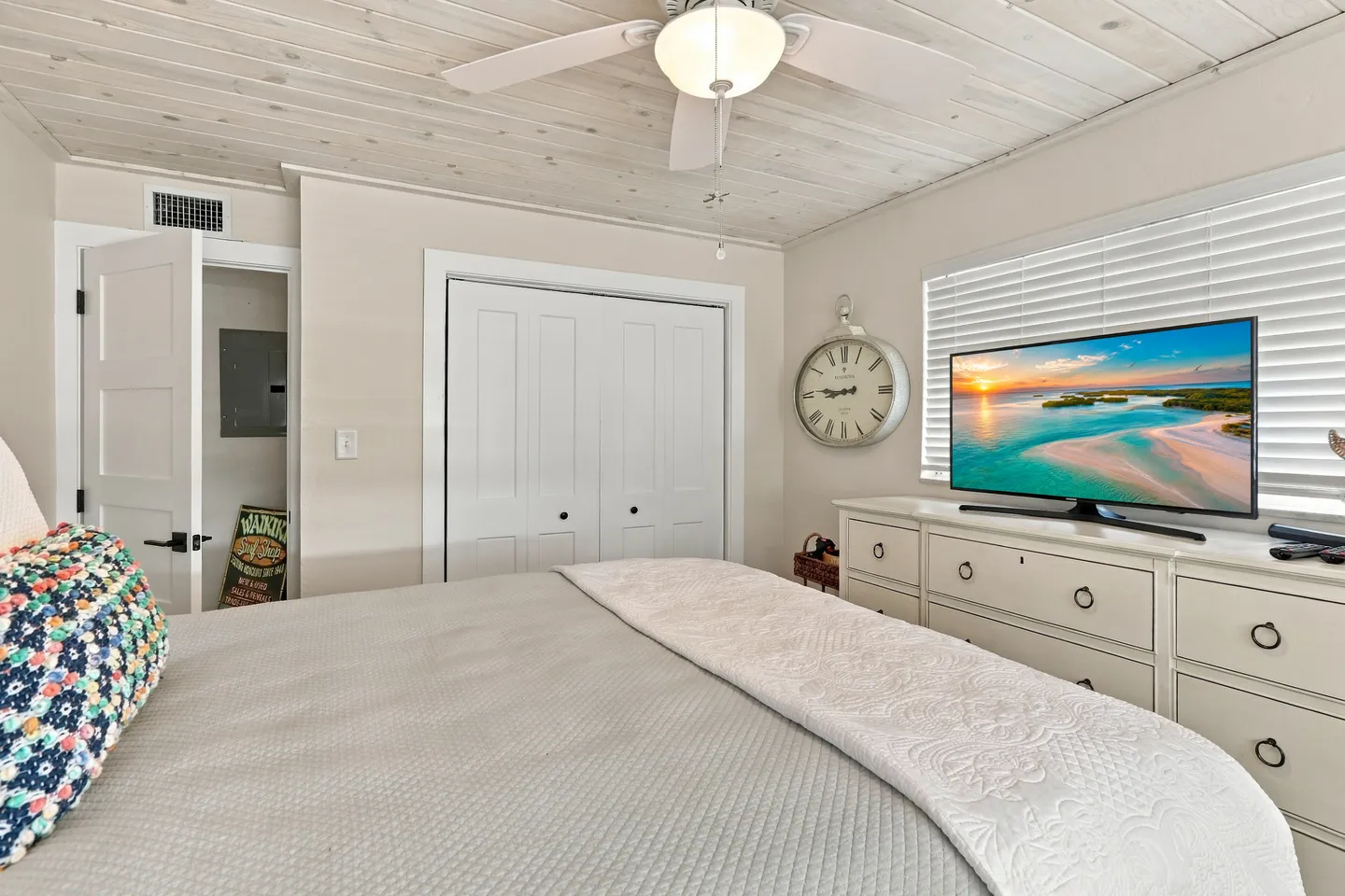 Bedroom with a bed in the foreground, white dresser with a flat-screen television, wood-paneled ceiling, and ceiling fan.