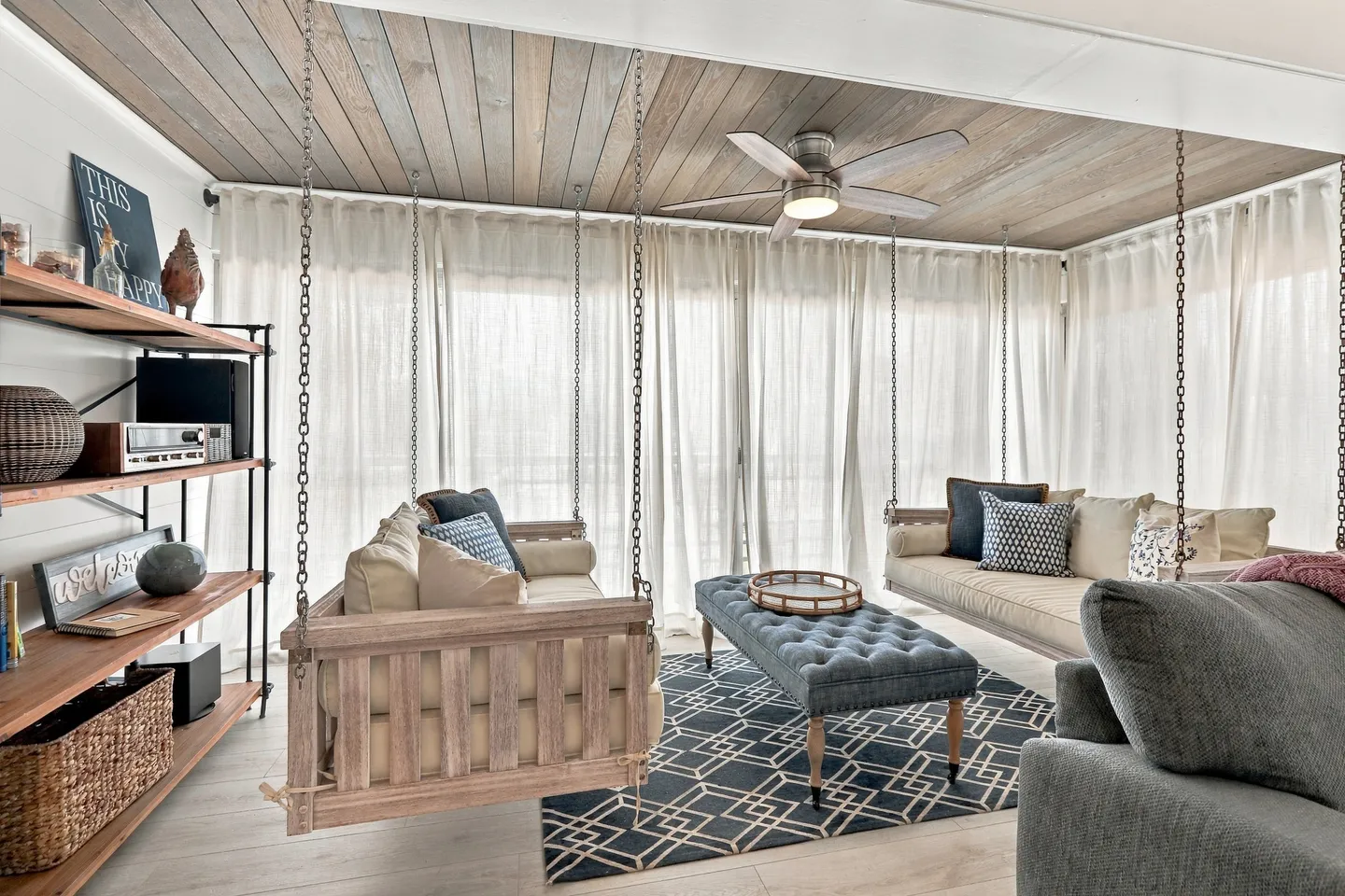 Sunroom seating area featuring two suspended wooden daybed swings, an upholstered ottoman, and sheer white curtains.