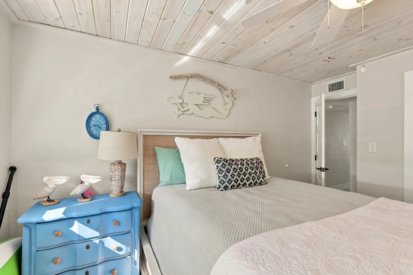 Bedroom featuring a bed with a woven headboard, a blue painted nightstand dresser with a lamp and bird figurines, and a whitewashed wood-plank ceiling.