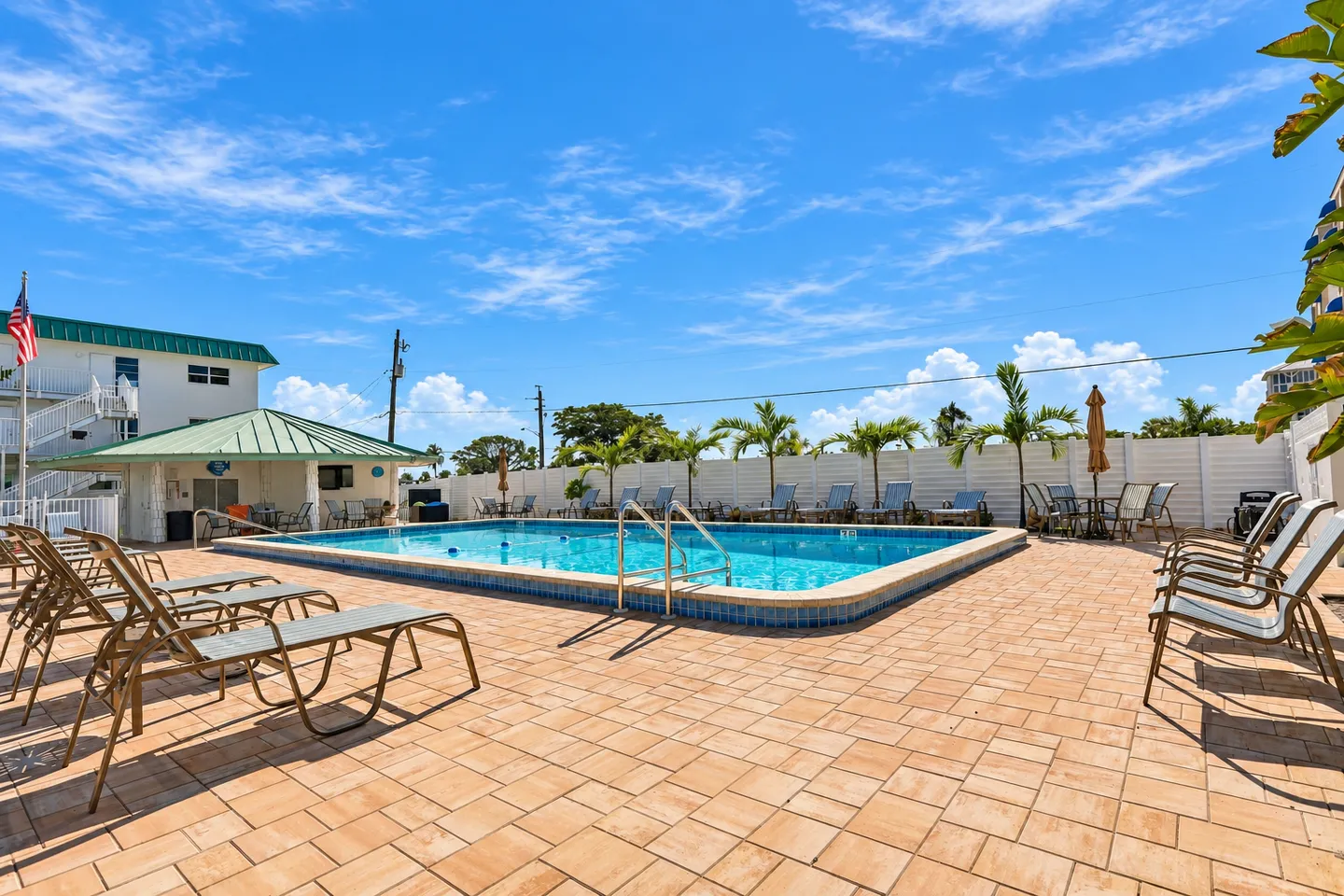 Outdoor swimming pool surrounded by a paved sun deck, lounge chairs, and a white fence under a blue sky.