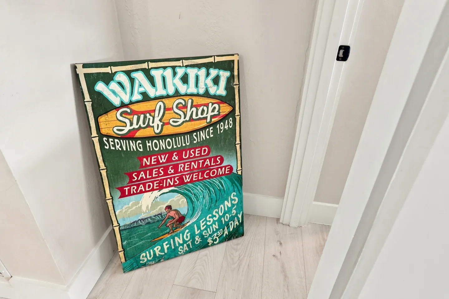 A vintage-style Waikiki Surf Shop decorative sign resting against an indoor wall on light wood-style flooring.