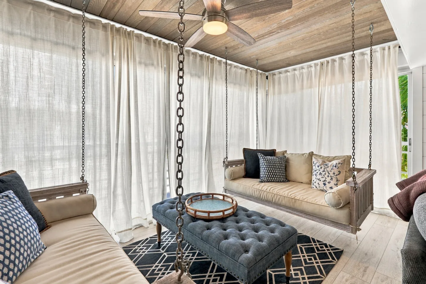 Porch area with suspended daybed swings, a tufted ottoman, and sheer curtains.