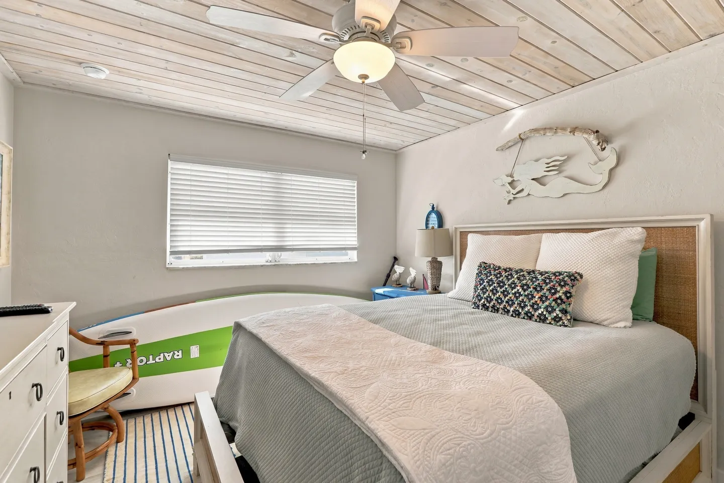 Bedroom featuring a made bed with a light blue quilt, a wood-paneled ceiling with a fan, a surfboard leaning against the wall, and a window with blinds.