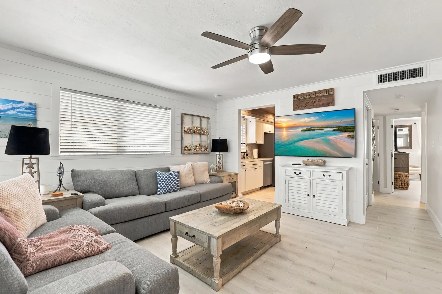 Bright living room with a gray sectional sofa, rustic wooden coffee table, ceiling fan, and wall-mounted TV above a white cabinet.