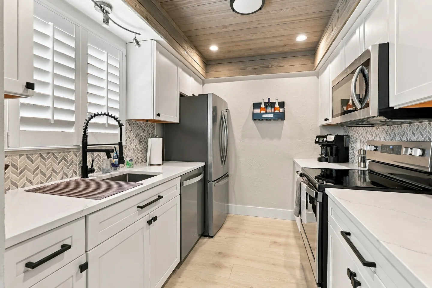 Galley-style kitchen with white cabinets, stainless steel appliances, herringbone backsplash, and a wood-plank ceiling.