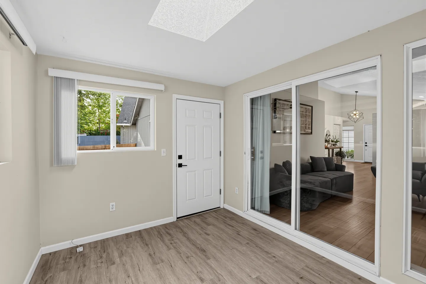 Enclosed entryway or sunroom with wood-style flooring, a white exterior door, a window with vertical blinds, and sliding glass doors looking into the living room.