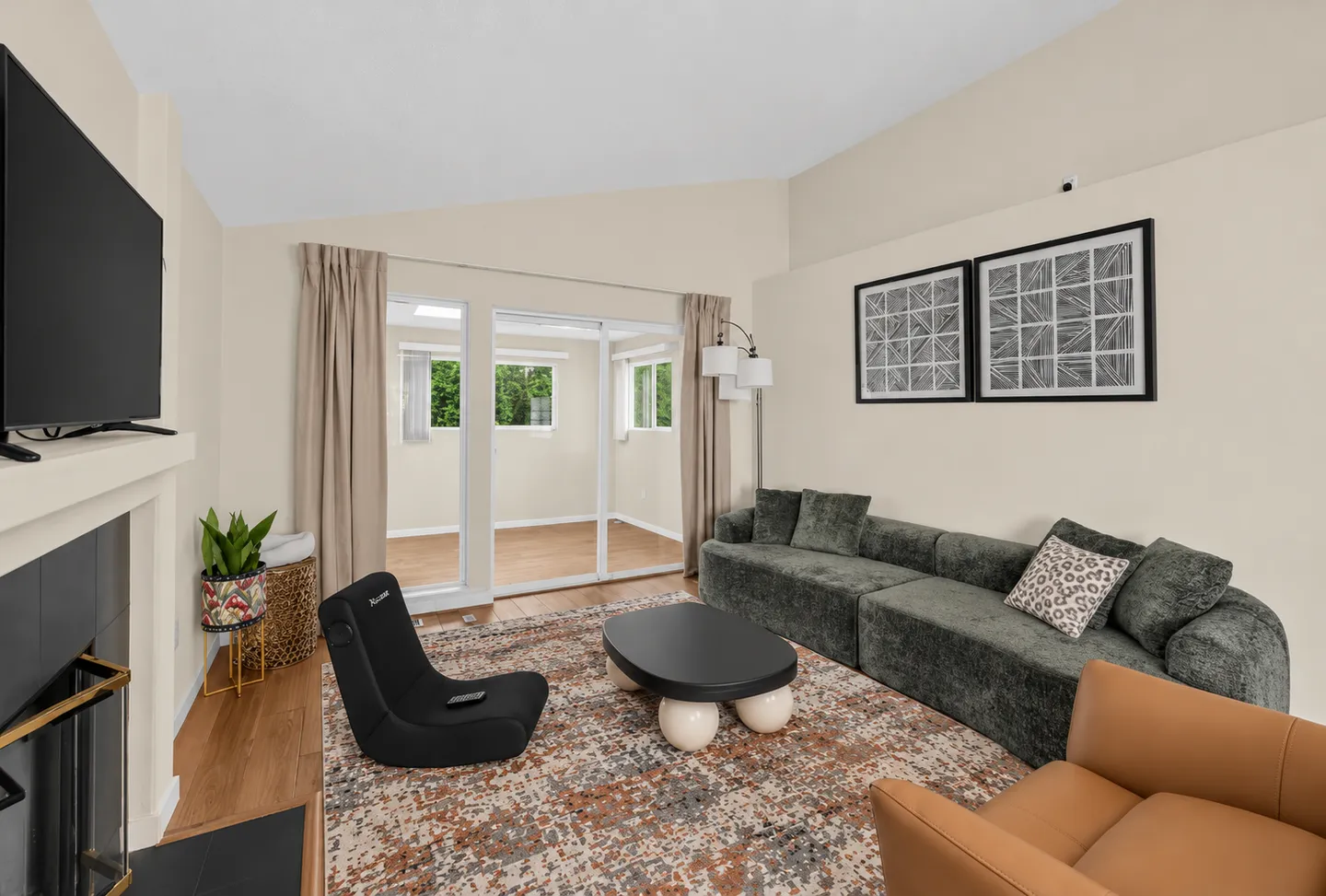 Living room featuring a grey sectional sofa, low black coffee table, modern floor chair, television above a fireplace, and glass sliding doors leading to an adjacent sunroom.
