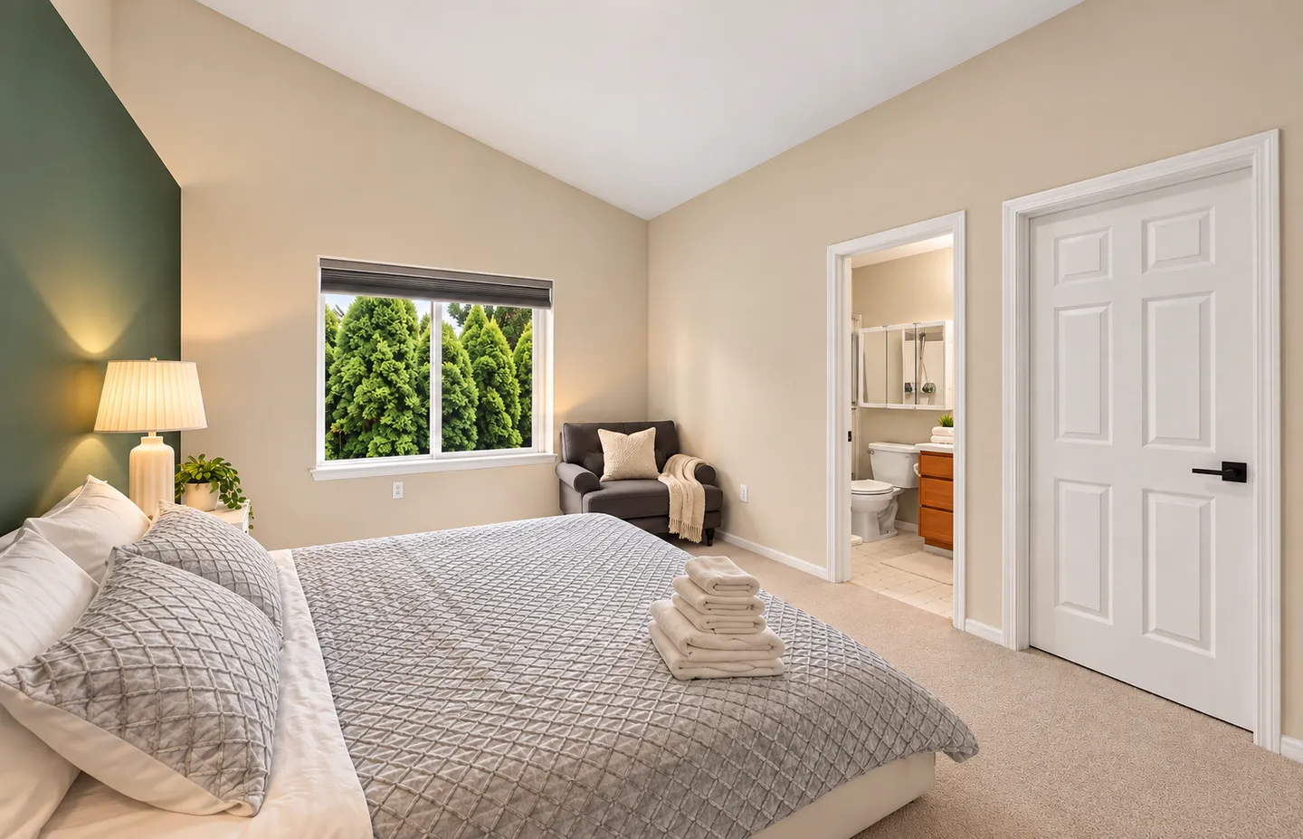 Bedroom with vaulted ceiling, made bed, accent armchair in the corner, and open doorway showing an en-suite bathroom.