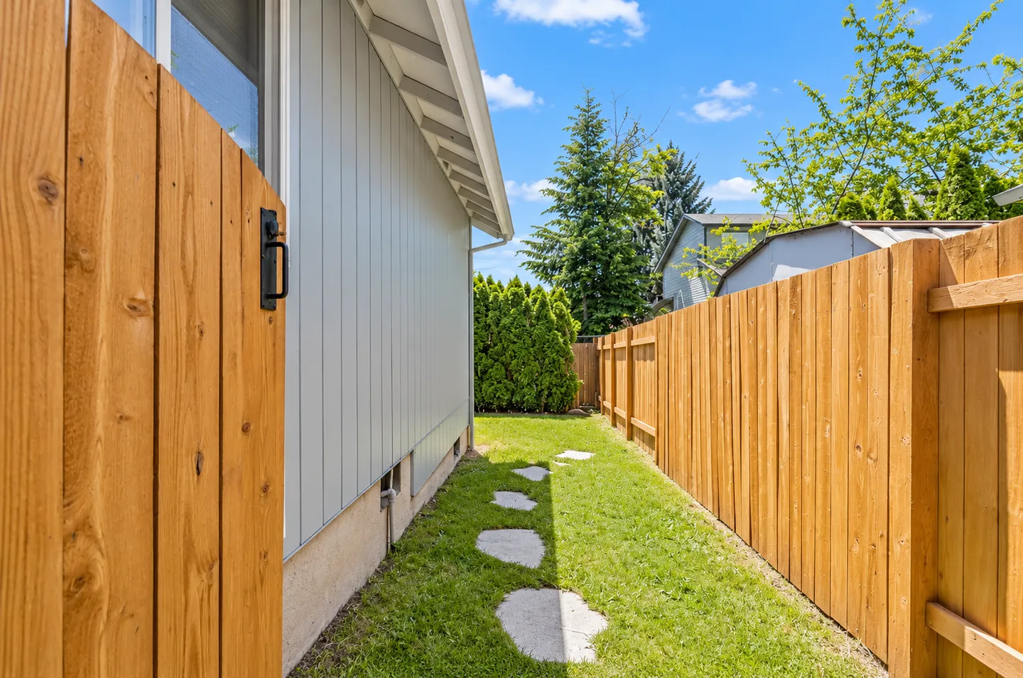 A narrow fenced side yard with green lawn, stone pavers, and a wooden gate along the house exterior.