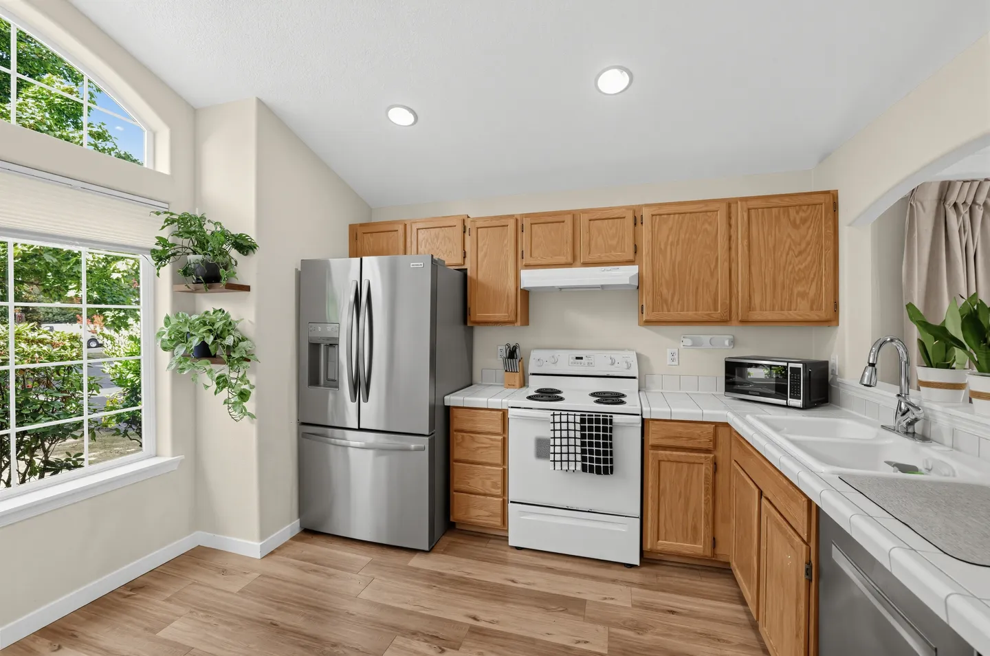 Kitchen with wood cabinetry, white tile countertops, stainless steel refrigerator, and large multi-pane windows.