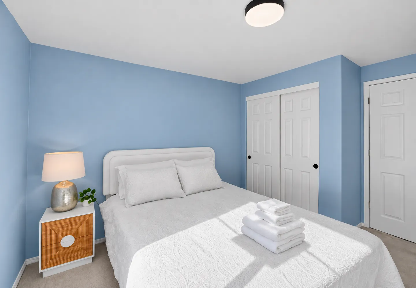 Bedroom with light blue walls, a bed made with white linens, a bedside table with a lamp, and white closet doors.