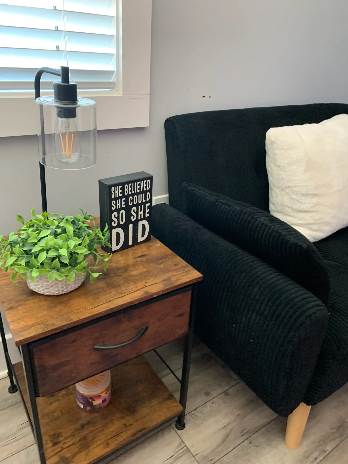 A wooden nightstand with a drawer beside a dark corduroy sofa with a white pillow, featuring a table lamp, small plant, and a sign.