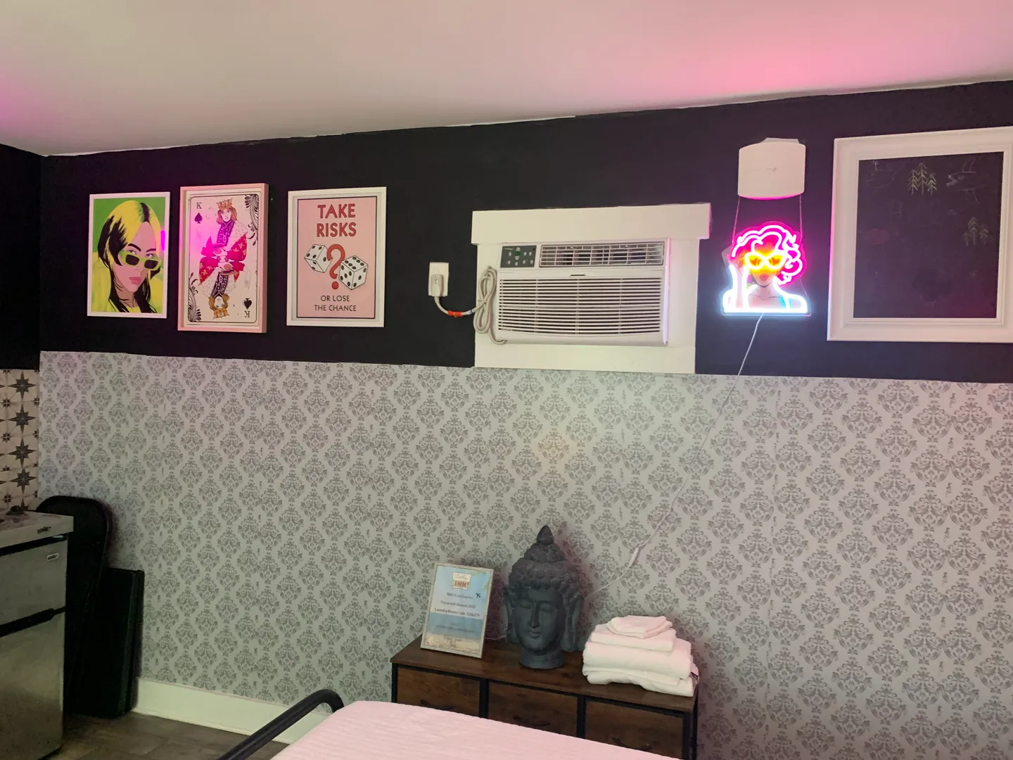 Bedroom wall with patterned wallpaper on the bottom, black paint on top, framed posters, a neon sign, a wall air conditioning unit, and a small dresser with folded towels and a Buddha head bust.