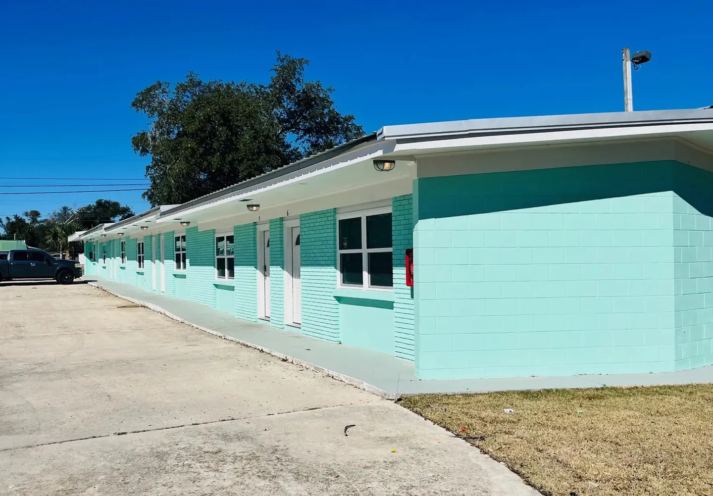 Exterior of a single-story motel-style building with turquoise painted brick walls, white doors, and a concrete driveway under a clear blue sky.