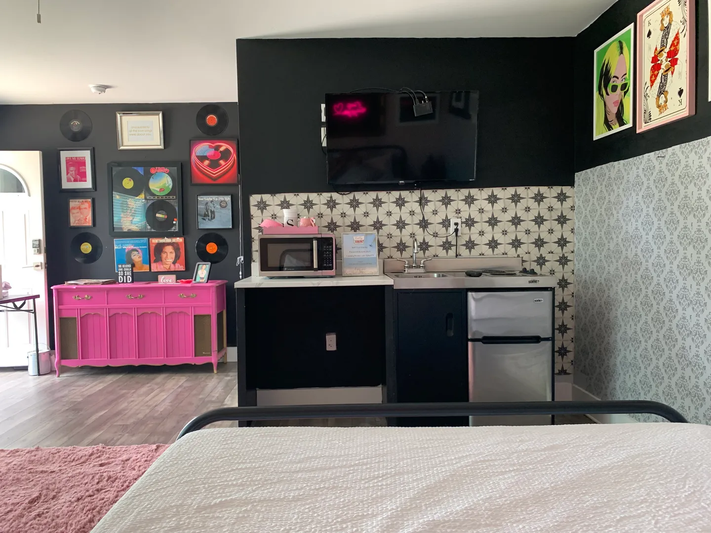 Bedroom kitchenette area with a mounted TV, compact sink and mini-fridge setup, microwave, and a bright pink sideboard beneath vinyl record wall decor.