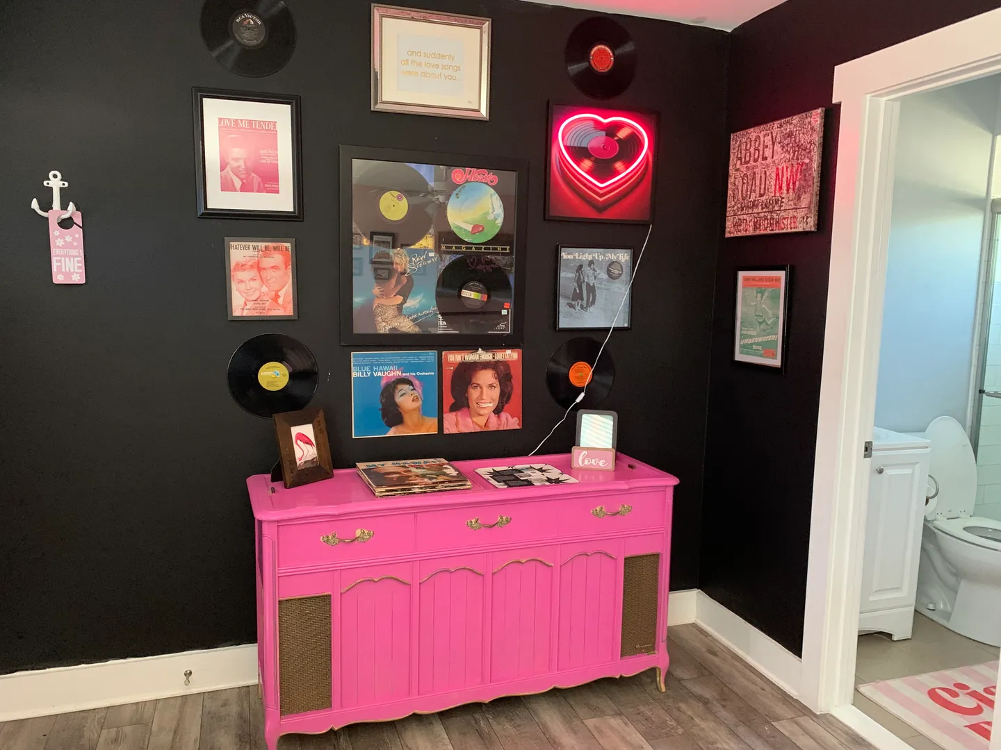 Bright pink vintage console cabinet against a black wall decorated with vinyl records and music-themed framed art next to an open bathroom door.