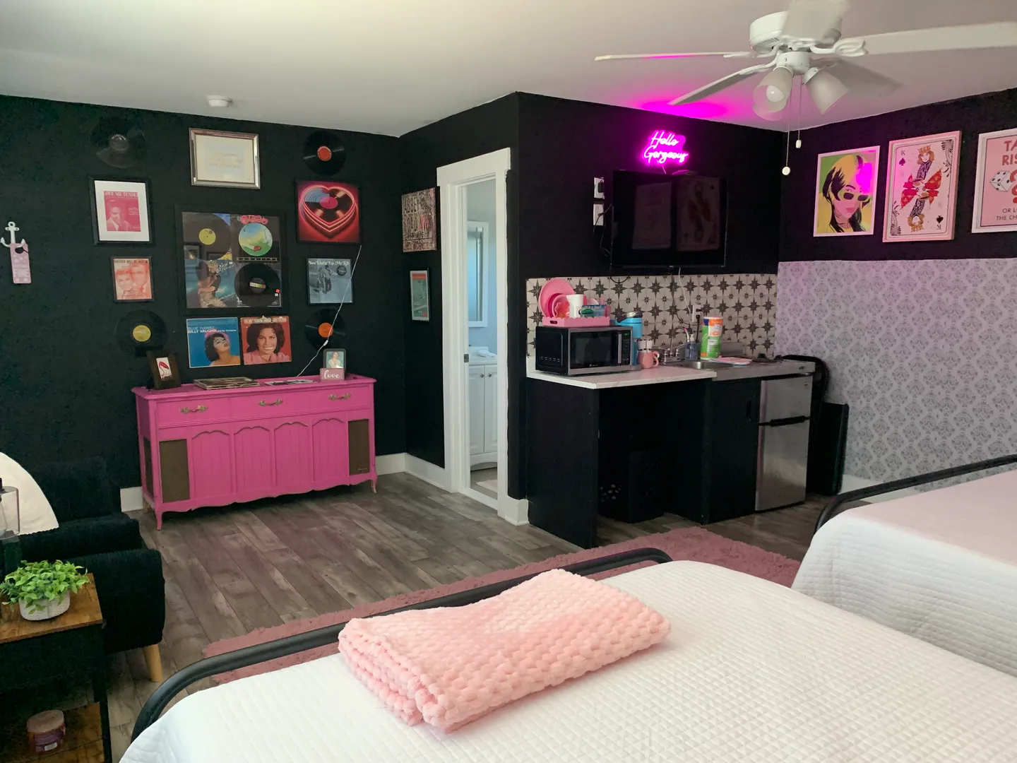 Bedroom interior featuring black accent walls decorated with retro vinyl records, a bright pink credenza, a kitchenette corner, and beds with white bedding.