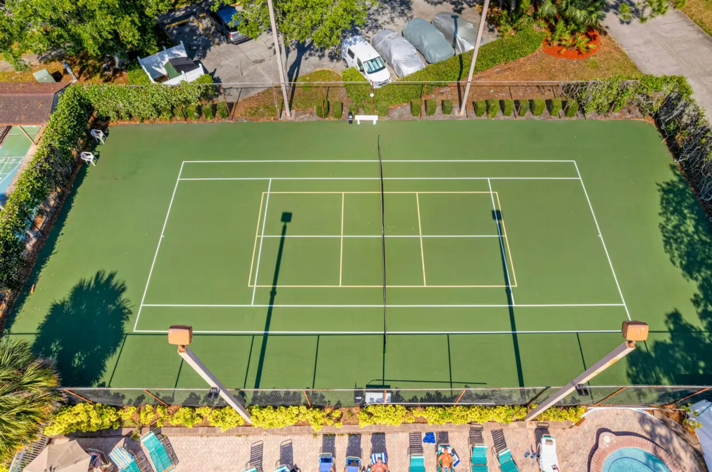 Elevated view of an outdoor green tennis and pickleball court enclosed by fencing, bordered by tropical landscaping and a pool deck.
