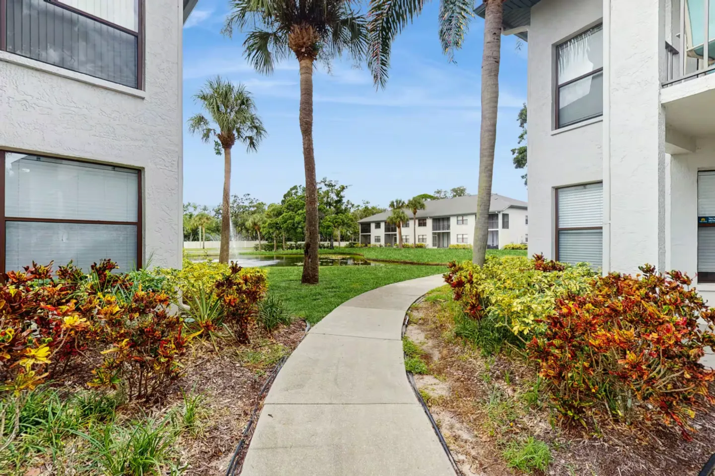 A concrete walkway winds between two apartment buildings toward a pond with a water fountain and palm trees.