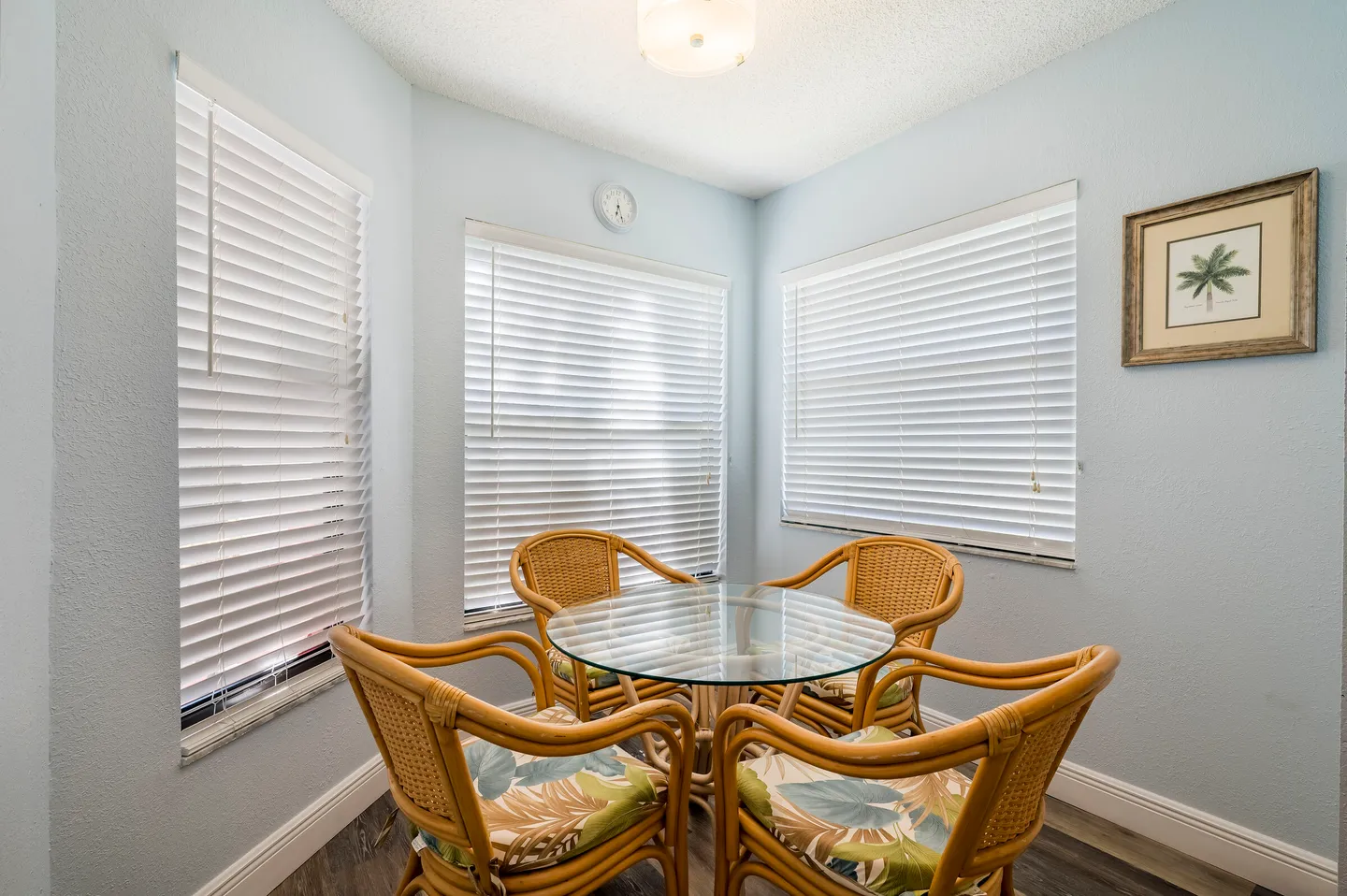 A breakfast nook featuring a round glass-top table surrounded by four wicker chairs with tropical-patterned cushions in front of three shuttered windows.