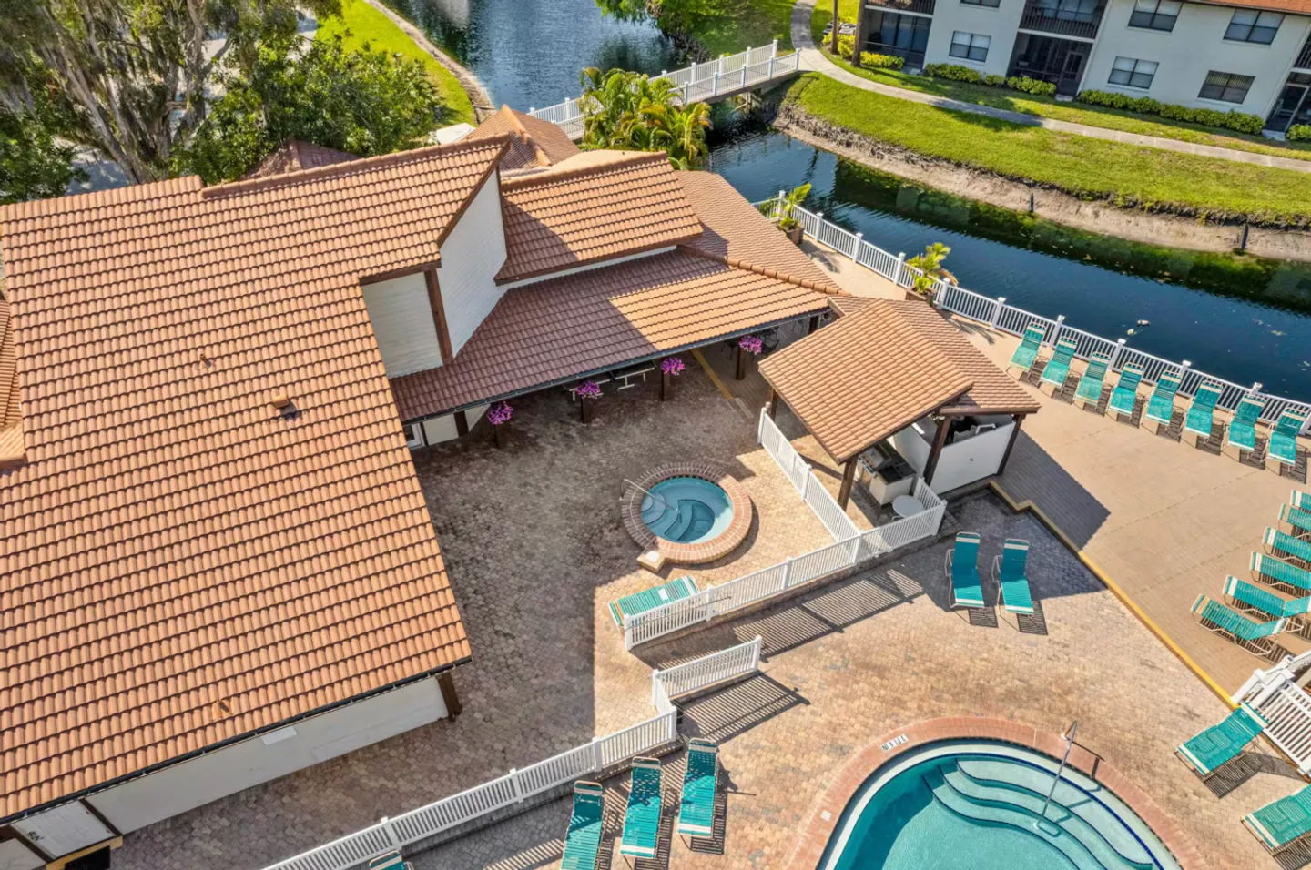 Aerial view of an outdoor community pool deck featuring a hot tub, lounge chairs, covered pavilion, and canal view.